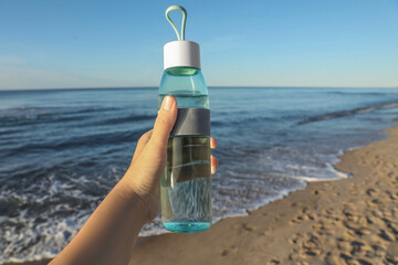 Fototapeta na wymiar Woman holding glass bottle with water near sea, closeup