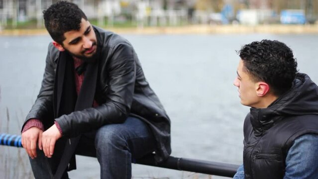 Two young middle eastern man talking to each other. They are located outside near a pond in a city