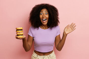 Photo of positive curly haired girl with surprised expression, raises palm, holds tasty doughnuts in one hand and looks directly to the camera with wide opened eyes, isolated over pink background. 