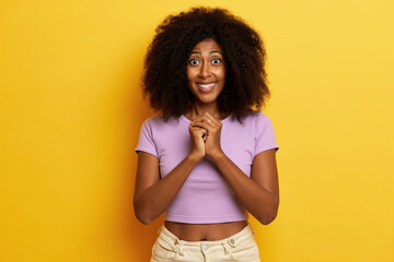 Horizontal shot of excited dark skinned model with curly hairstyle keeps hands pressed together under chin, smiles toothily with wide opened eyes, wears purple t-shirt and jeans, isolated over yellow