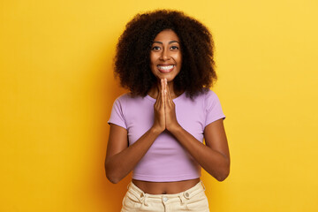 Positive charming curly haired woman holds palms pressed together and smiles widely, poses over yellow background