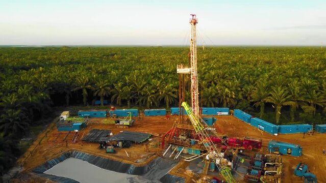 Cinematic 4K Drone Footage of Onshore Drilling Rig equipment structure for oil exploration and exploitation in the middle of jungle surrounded by palm oil trees during sunset and high oil price.