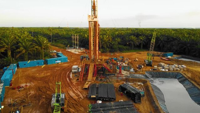 Cinematic 4K Drone Footage of Onshore Drilling Rig equipment structure for oil exploration and exploitation in the middle of jungle surrounded by palm oil trees during sunset and high oil price.
