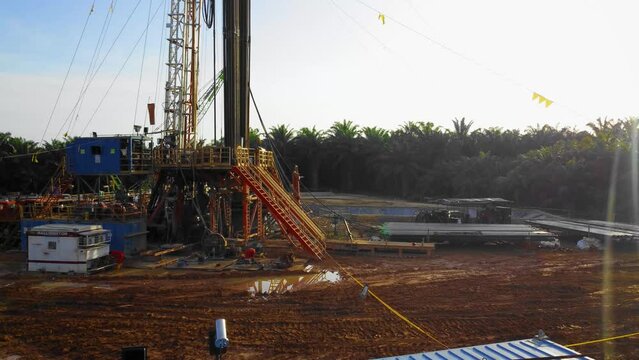Cinematic 4K Drone Footage of Onshore Drilling Rig equipment structure for oil exploration and exploitation in the middle of jungle surrounded by palm oil trees during sunset and high oil price.