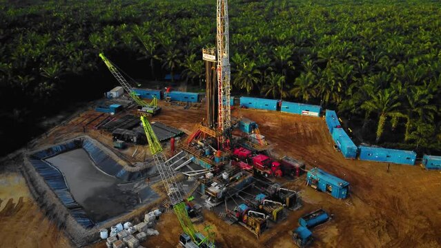 Cinematic 4K Drone Footage of Onshore Drilling Rig equipment structure for oil exploration and exploitation in the middle of jungle surrounded by palm oil trees during sunset and high oil price.