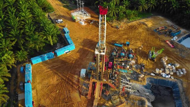 Cinematic 4K Drone Footage of Onshore Drilling Rig equipment structure for oil exploration and exploitation in the middle of jungle surrounded by palm oil trees during sunset and high oil price.