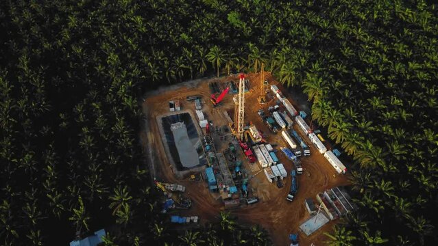 Cinematic 4K Drone Footage of Onshore Drilling Rig equipment structure for oil exploration and exploitation in the middle of jungle surrounded by palm oil trees during sunset and high oil price.