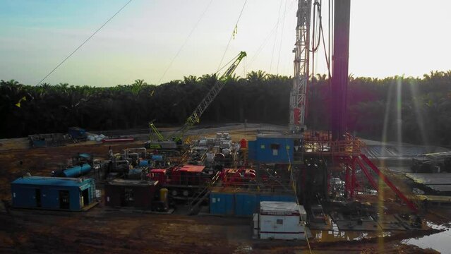 Cinematic 4K Drone Footage of Onshore Drilling Rig equipment structure for oil exploration and exploitation in the middle of jungle surrounded by palm oil trees during sunset and high oil price.