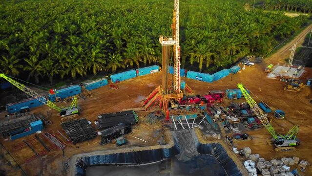 Cinematic 4K Drone Footage of Onshore Drilling Rig equipment structure for oil exploration and exploitation in the middle of jungle surrounded by palm oil trees during sunset and high oil price.