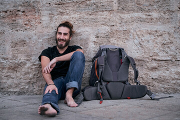 caucasian guy with beard long hair barefoot sitting on the floor leaning against the wall smiling with dirty feet next to his backpack