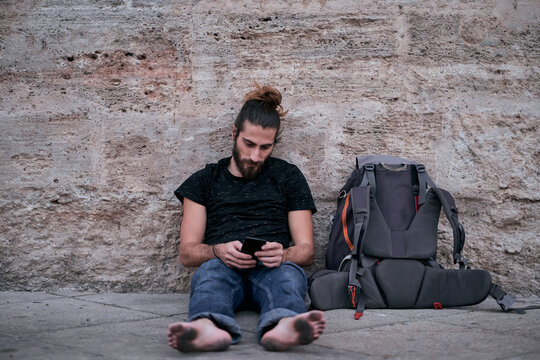 caucasian guy with beard long hair barefoot looking and touching his smartphone sitting on the floor leaning against the wall with his dirty feet next to his backpack