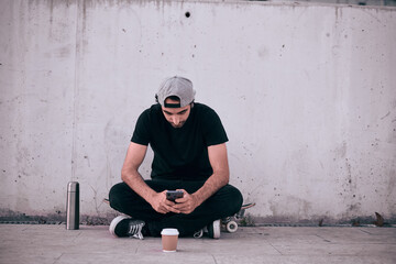 caucasian guy in a gray cap and black t-shirt sitting on his skateboard manipulating his smartphone next to a thermos and a glass of coffee