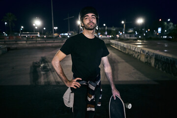 caucasian guy in a striped sweater tied on the waist holding a skateboard with his hand looking at camera in the city night