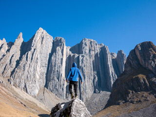 Fototapeta premium Hombre parado sobre una roca o piedra, con una casaca o chamarra color azul, de espalda observando una gran montaña de rocas, y el cielo azul despejado, en Perú Sudamérica