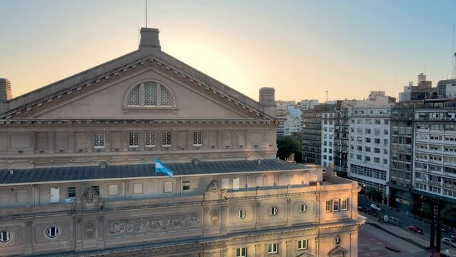 Sunset view, aerial reverse flyover 9 de julio avenue with busy traffics, overlooking at the rear entrance of iconic landmark teatro colon performing theatre at downtown Buenos Aires city, Argentina.