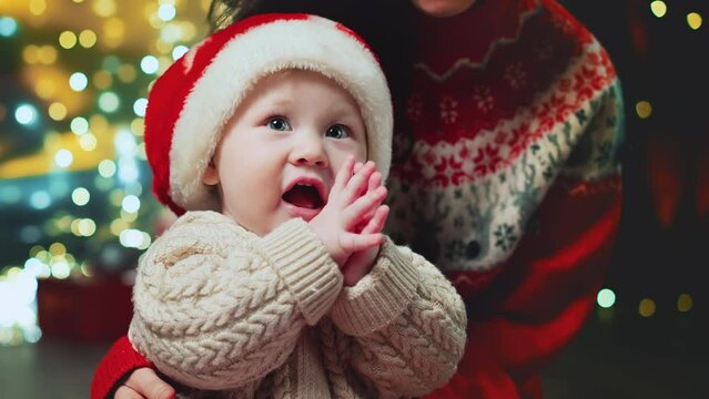 Sweet little asian baby boy and Mother at background of decorated fir-tree play with santa hat of Christmas. Happy mom unpack present or gift. Merry Christmas, happy new year toddler clapping hands