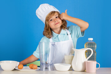 Portrait of a 7, 8 years old child in cook cap and apron making fruit salad and cooking food in kitchen. Cute little blonde happy smiling chef.