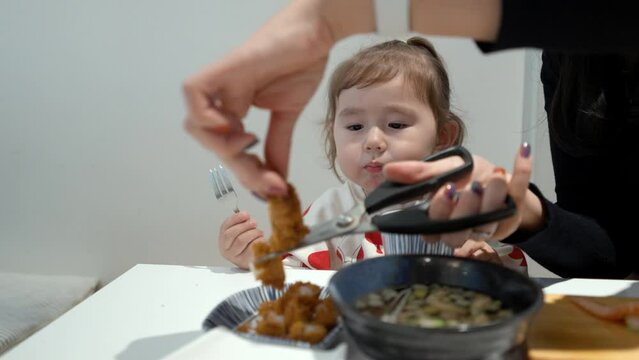 Mother's Hands Cutting Fried Fish Fillet Into Bite Size Pieces. Beautiful Female Toddler Munching Food While Watching. medium shot