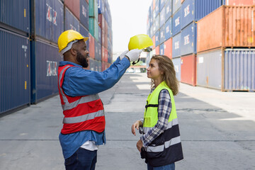 A  foreman with mustache and beard helps a female apprentice wear a hardhat. Both of them wearing reflective safety vest. There are container in the work area.