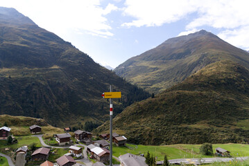 Fototapeta na wymiar Hiking trail sign with aerial view of mountain village Tschamut in the Swiss Alps at region of Oberalppass on a blue cloudy late summer day. Photo taken September 5th, 2022, Oberalp Pass, Switzerland.