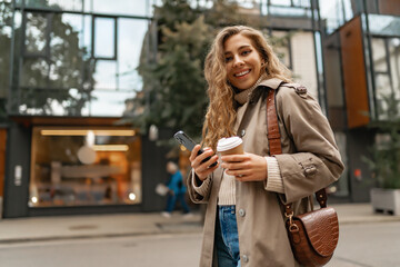 Young woman with curly blonde hair using the phone with a cup of coffee in hands on the city streets