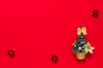 This small decorative artificial Christmas tree is adorned with golden bow and flowers. On a red background with a place for text or congratulations are New Year's decor items: spruce and toy balls