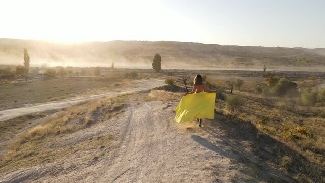 Drone footage back view of girl walking in a long yellow dress in the mountains of Cappadocia at sunset