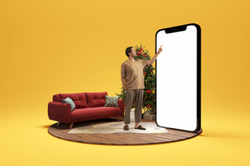 Christmas atmosphere at home. Photo and 3d illustration of man standing next to huge 3d model of smartphone with empty white screen isolated on yellow background. Mockup for ad - obrazy, fototapety, plakaty