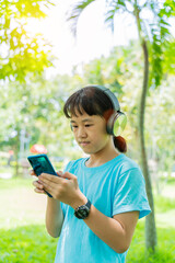 Pretty school girl with smartphone and listens to music with modern headphones wireless in park outdoors.