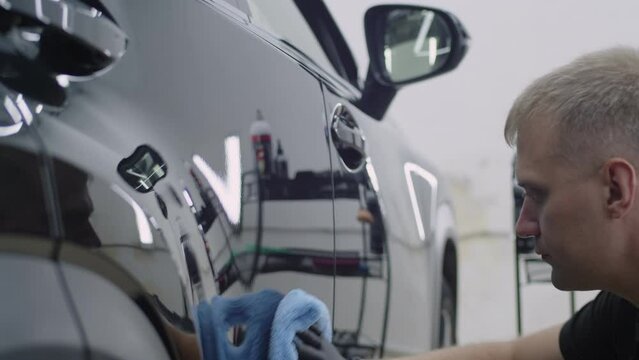 Close-up. A man professionally makes car detailing in a car service. Caucasian guy in a black glove wipes the surface of a black car with a rag until it shines.