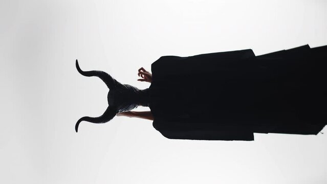 Silhouette of a beautiful woman in a maleficent costume. Woman seduces with mystical hand gestures. A girl in a black devilish costume with horns on a white background. Vertical video.