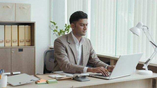 Indian businessman typing on laptop and reading papers while working at desk in office