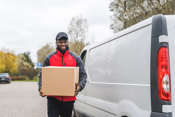 Black handsome delivery-person in red-and-gray jacket and black hat carrying one cardboard box....