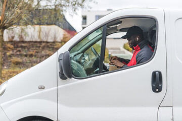 Postal ethnic male courier inside white delivery van sitting on driver's seat and woking on his laptop. Autumn weather. Package delivery concept. High quality photo