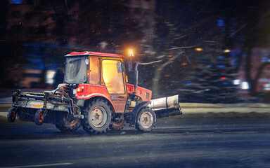 Tractor with snow plow and rear brush for sweeping sidewalks, walkways and roads moving on city road at night during snow storm, road maintenance at winter season. Tractor cleaning snow and ice - obrazy, fototapety, plakaty