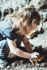 Beautiful young boy child with long blond hair playing construction site in the dirt with diggers and dumpers while out in nature. Free and independent play while hiking in a natural park.