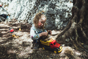 Beautiful young boy child with long blond hair playing construction site in the dirt with diggers and dumpers while out in nature. Free and independent play while hiking in a natural park.