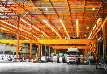 Front view of truck loading steel coil under overhead crane inside orange color warehouse. Steel industry handling and transportation concept.
