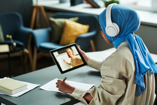 Young Muslim woman in blue hijab and white headphones listening to explanation of teacher during online lesson and making notes