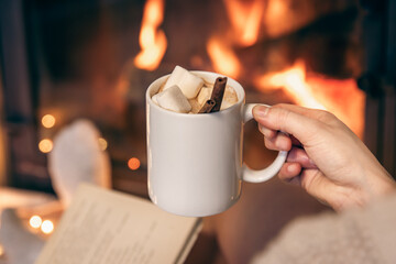 A cup of cocoa with marshmallows in female hands by the fireplace.