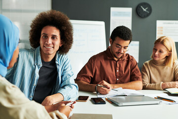 Happy young man with thick dark hair looking at Muslim female colleague in blue hijab during conversation by workplace