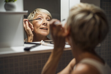 Mature woman is applying mascara in the bathroom.