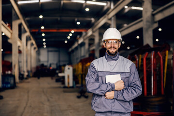 A factory supervisor in work wear is holding tablet and smiling at the camera in facility.