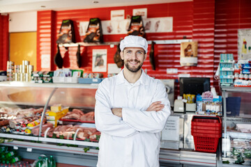 A butchery salesman stands in the butchery with arms crossed and smiles at the camera.