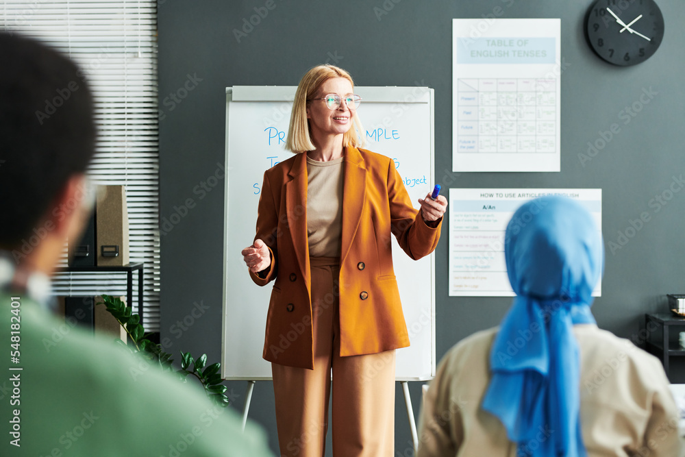 Poster happy mature blond teacher or speaker standing in front of audience and explaining them new grammar 