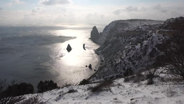 Snow covered rocky cliffs over sea. Winter landscape over the sea bay. Juniper tree under snow. Concept of winter holidays, travel and recreation. Cape Fiolent in Balaklava, Sevastopol, Crimea