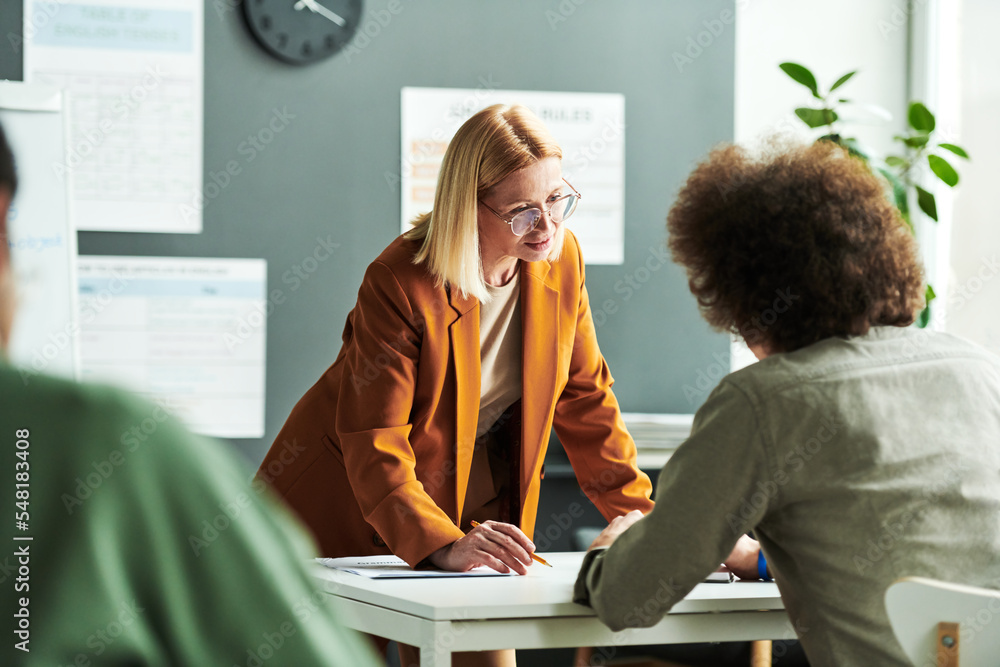 Poster Blond mature teacher of foreign language communicating with student at lesson while standing in front of him during consultation