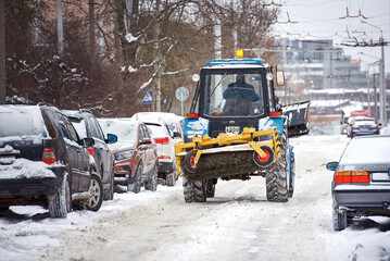 Tractor with snow plow and rear brush removing snow from road during snow storm, road maintenance at winter season. Tractor clean road and remove snow and ice from parking lot