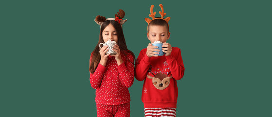 Cute children in Christmas pajamas drinking hot chocolate on green background