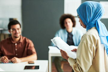 Young Muslim teacher in blue hijab looking at text in document while making report or explaining new subject to students